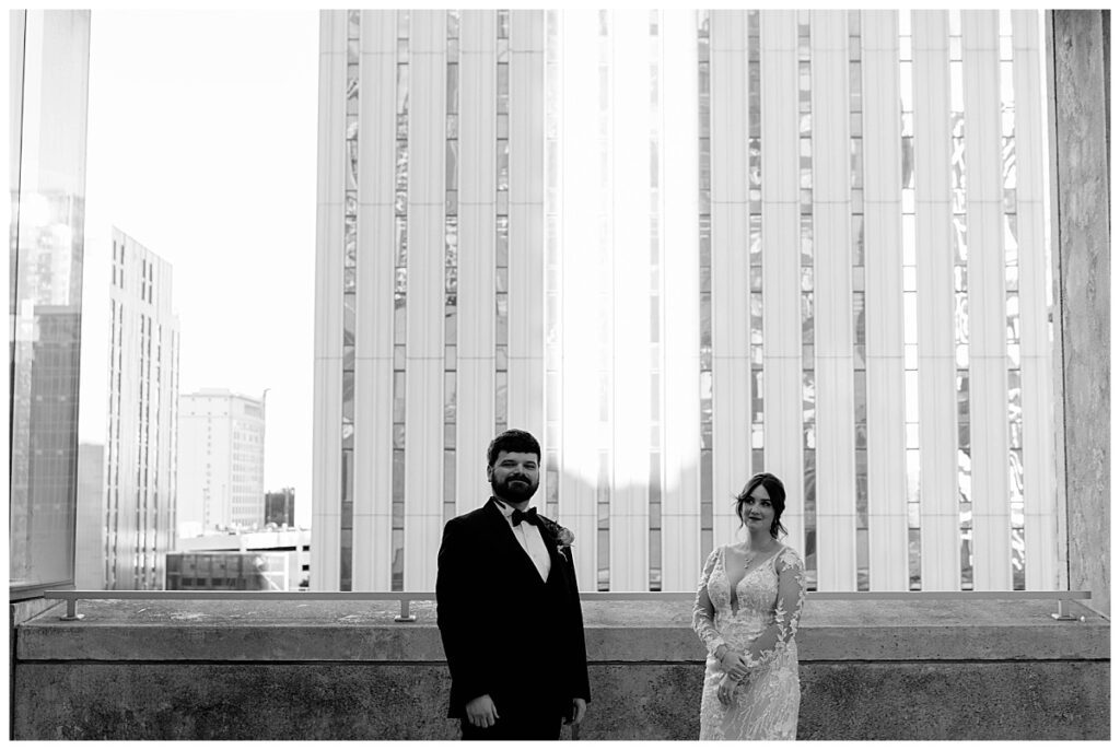 man and woman stand on balcony overlooking city buildings by The Hazel Club