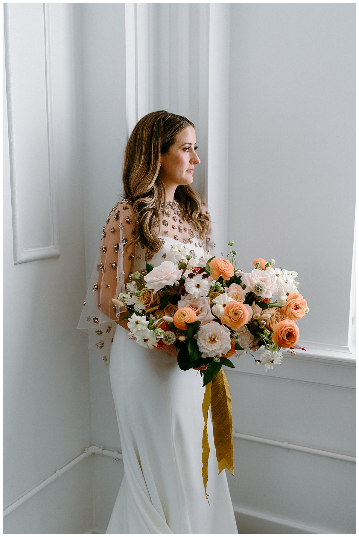 bride wearing cape holds bouquet and looks out window by The Hazel Club