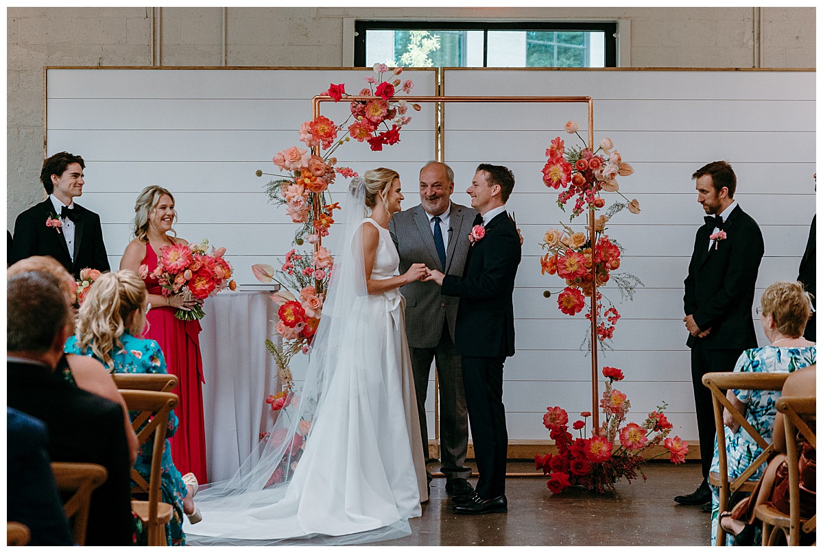 couple stands together under ceremony arch holding hands by Charlotte wedding photographer